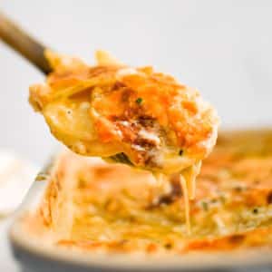 Large wooden serving spoon filled with gooey, cheesy, perfectly baked scalloped potatoes in a cream sauce and topped with cheese.