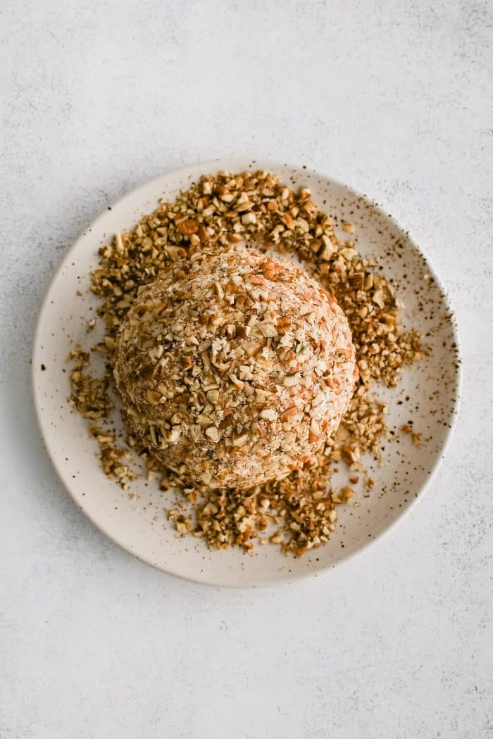 Refrigerated cheese ball coated in crushed pecans on a plate filled with crushed pecans.