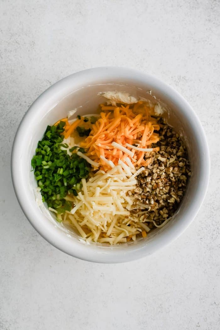 Chopped green onions, shredded cheddar cheese, shredded Monterey Jack cheese, and chopped pecans added to a white mixing bowl filled with softened cream cheese seasoned with garlic and onion powder.