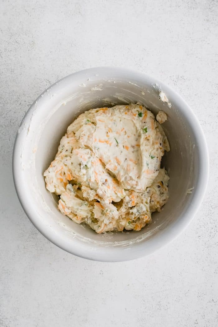 Cheese ball mixture in a large white mixing bowl consisting of cream cheese, chopped green onions, shredded cheddar cheese, shredded Monterey Jack cheese, and chopped pecans.