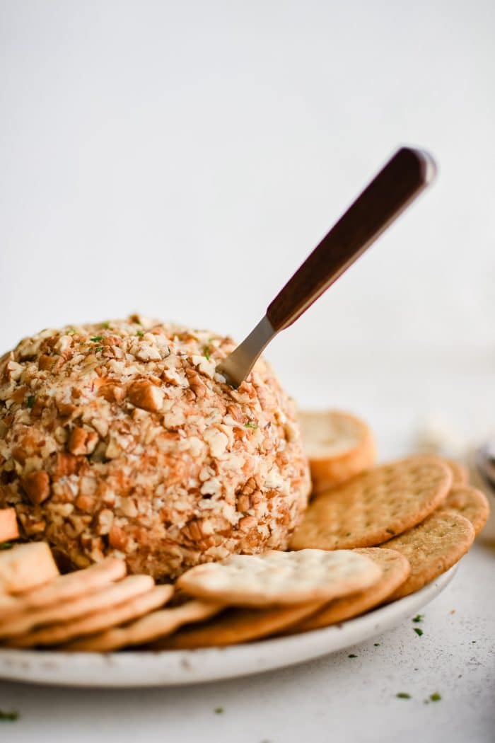 Classic cheese ball coated in chopped pecans and garnished with fresh parsley on a plate with a small spreader knife inserted in the side and served with various types of crackers and crostini.