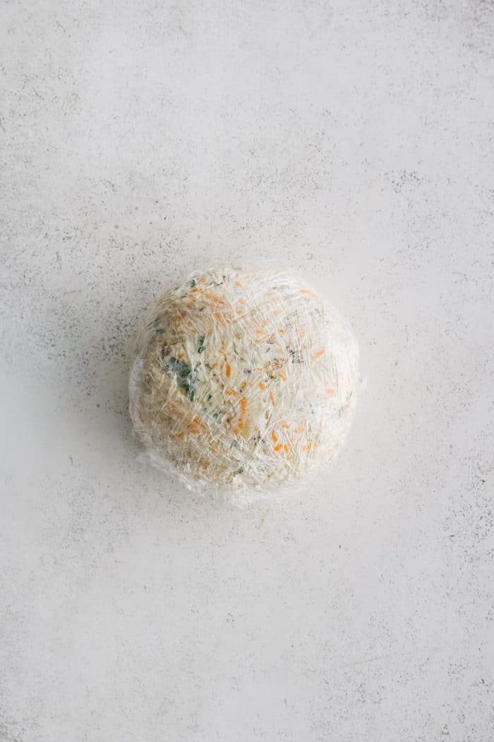 Cheese ball mixture shaped into a ball and wrapped twice with plastic wrap.