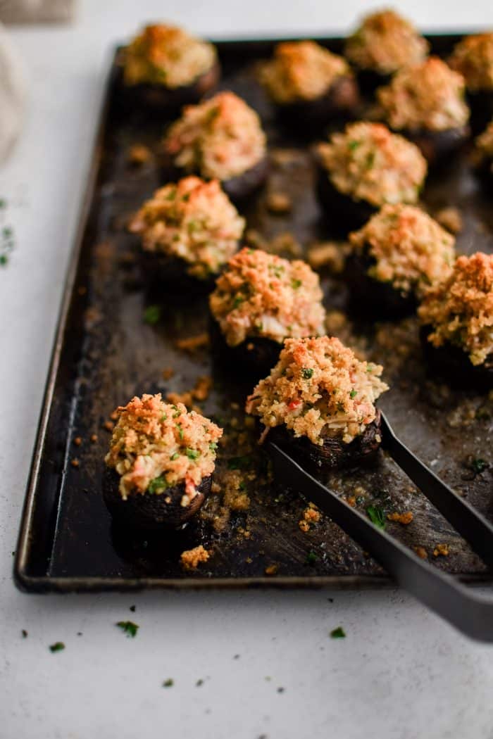 Small serving tongs removing one crab stuffed mushroom from a baking sheet.