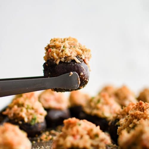 Small serving tongs holding one crab stuffed mushroom over a baking sheet with the remaining baked crab stuffed mushrooms.