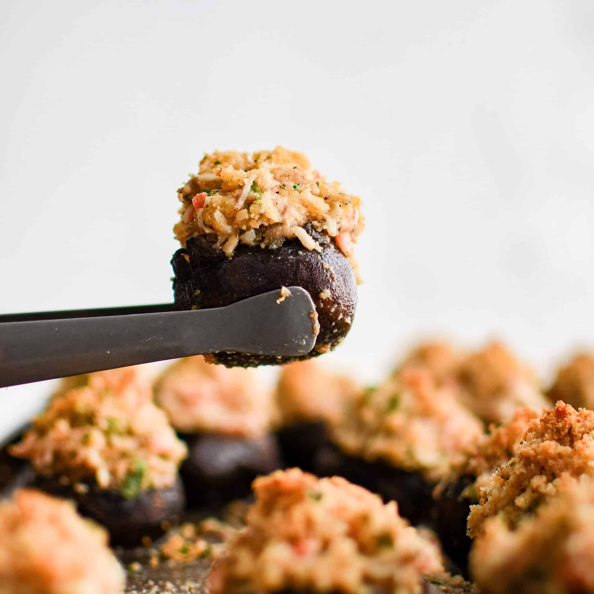 Small serving tongs holding one crab stuffed mushroom over a baking sheet with the remaining baked crab stuffed mushrooms.