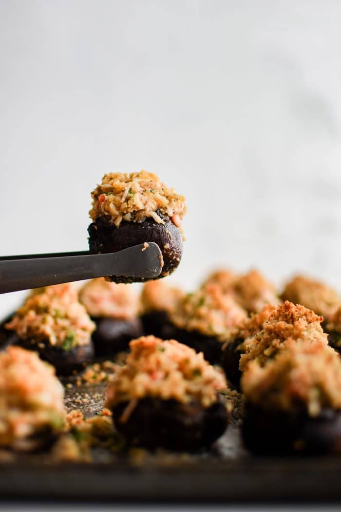 Small serving tongs holding one crab stuffed mushroom over a baking sheet with the remaining baked crab stuffed mushrooms.