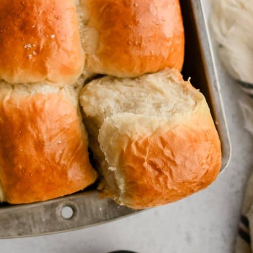 One corner dinner roll removed partially from a pan filled with perfectly baked yeast dinner rolls with golden tops.