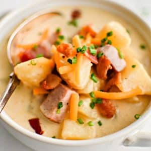 White soup bowl filled with chunks of potatoes and diced ham in a creamy broth garnished with fresh chives, freshly shredded cheddar cheese, and bacon bits.