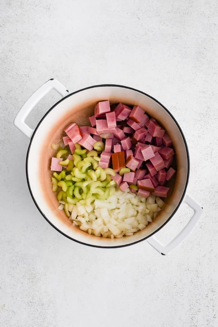 Large white ceramic Dutch oven filled with chunks of ham steak, diced onion, and diced celery cooking in melted butter.