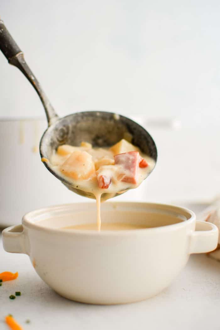 Large soup ladle pouring creamy ham and potato soup into a white soup bowl.