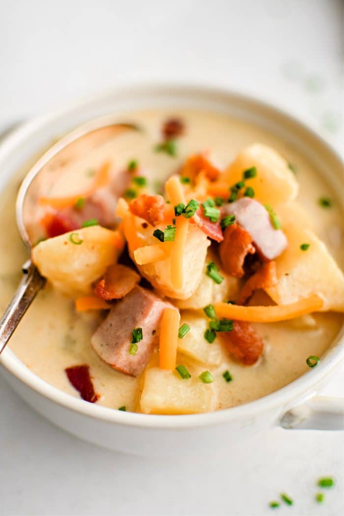 White soup bowl filled with chunks of potatoes and diced ham in a creamy broth garnished with fresh chives, freshly shredded cheddar cheese, and bacon bits.