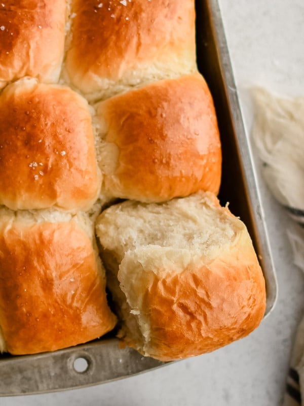 One corner dinner roll removed partially from a pan filled with perfectly baked yeast dinner rolls with golden tops.