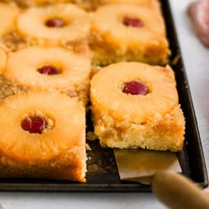 A metal spatula removing a single slice of pineapple upside down cake topped with one pineapple ring and one maraschino cherry.