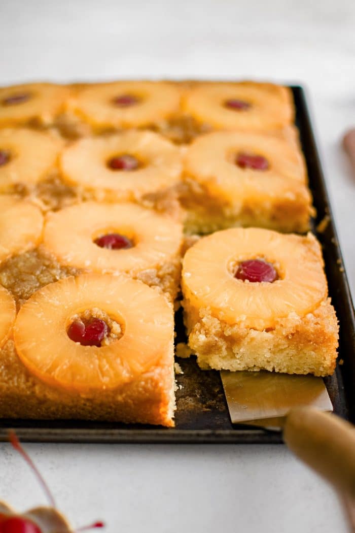 A metal spatula removing a single slice of pineapple upside down cake topped with one pineapple ring and one maraschino cherry.