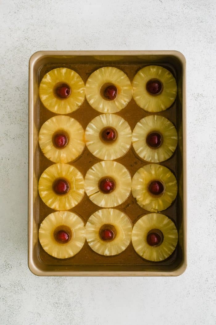 A 9x13 inch baking pan filled with melted butter mixed with brown sugar and topped with four rows of pineapple rings with maraschino cherries in the middle.