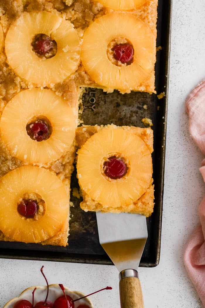 A metal spatula removing a single slice of pineapple upside down cake topped with one pineapple ring and one maraschino cherry.