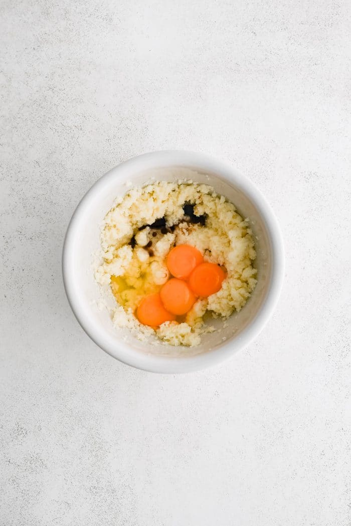 Vanilla extract, eggs, and egg yolk added to a mixing bowl filled with whipped butter and sugar.