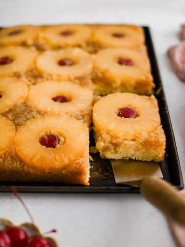 A metal spatula removing a single slice of pineapple upside down cake topped with one pineapple ring and one maraschino cherry.