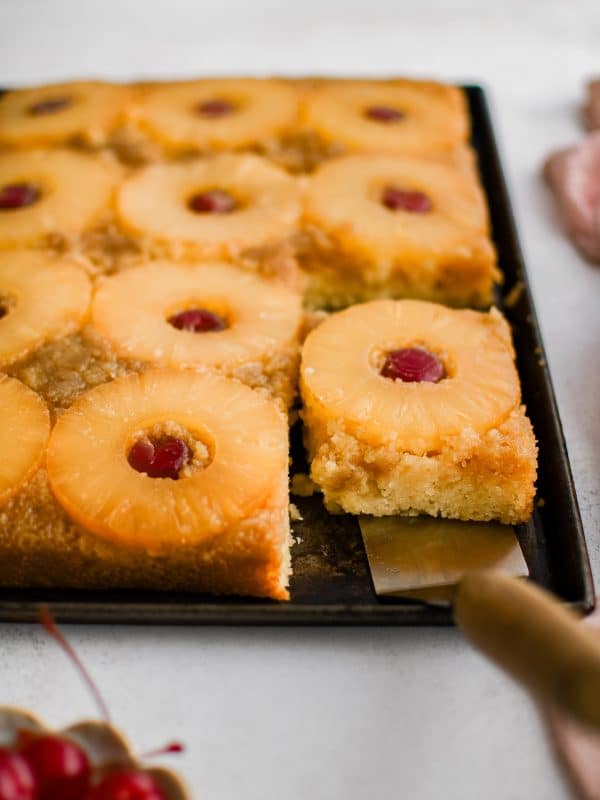 A metal spatula removing a single slice of pineapple upside down cake topped with one pineapple ring and one maraschino cherry.