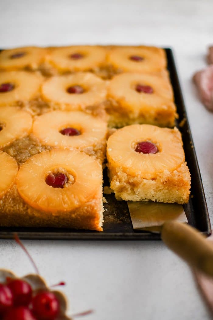 A metal spatula removing a single slice of pineapple upside down cake topped with one pineapple ring and one maraschino cherry.