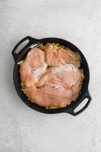 Large cast iron skillet filled with a bottom layer of crushed potato chips topped with thinly sliced raw chicken breasts.