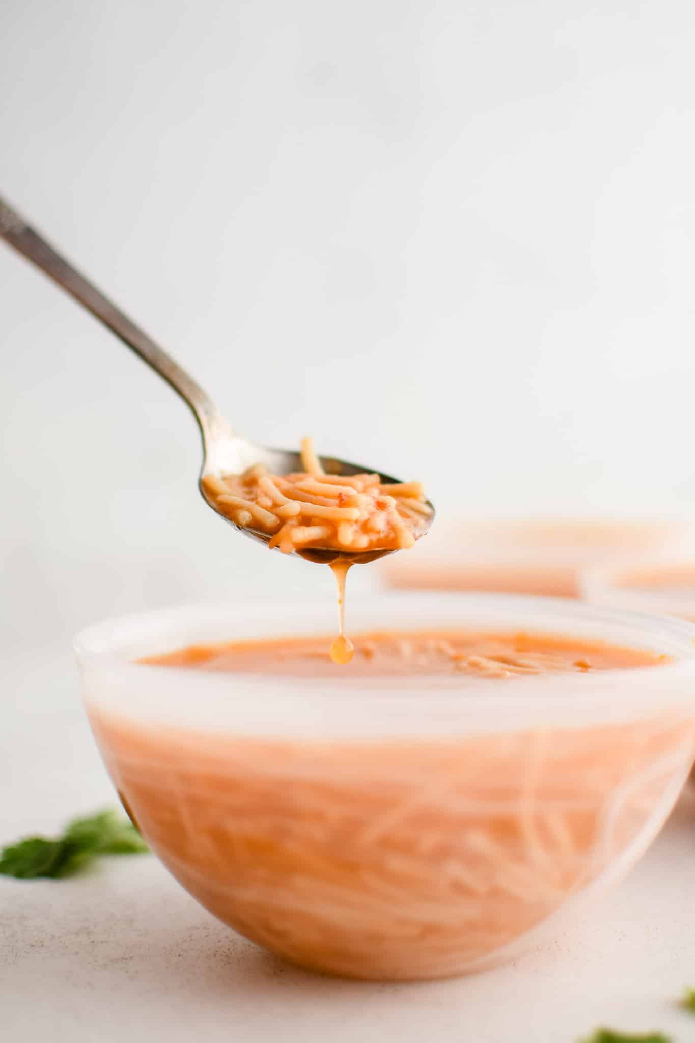 Spoon filled with soup hovering above a white, semi-transparent soup bowl filled with Sopa De Fideo (Mexican Noodle Soup) made with a light tomato broth and small fideo noodles.