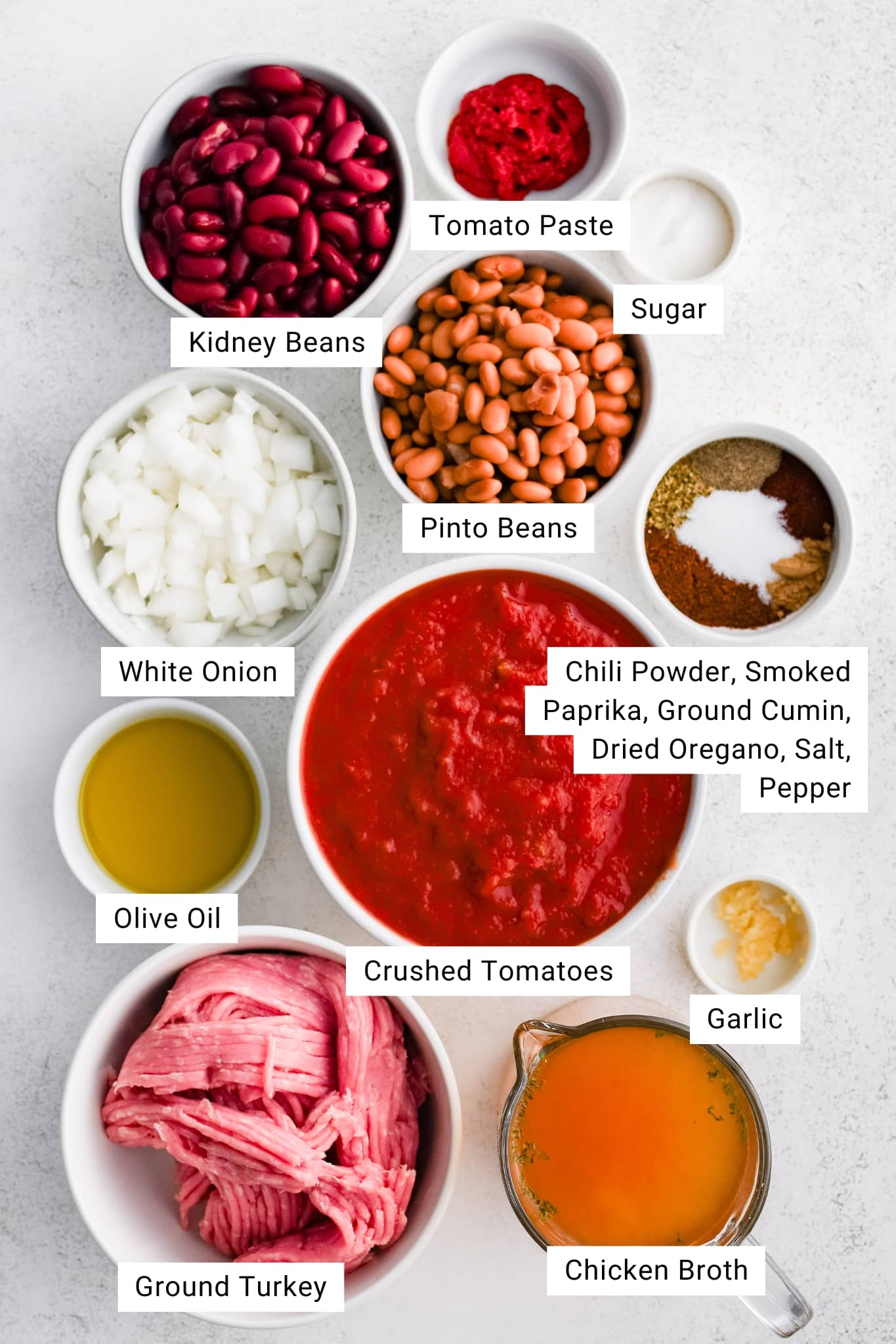 A flat lay of labeled ingredients for ground turkey chili arranged on a white surface. The photo features kidney beans, pinto beans, white onion, tomato paste, sugar, olive oil, crushed tomatoes, ground turkey, chicken broth, minced garlic, and a spice mix of chili powder, smoked paprika, cumin, oregano, salt, and pepper.