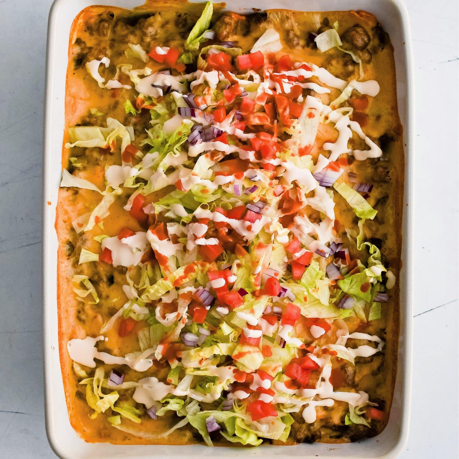Baked walking taco casserole in a large 9x13-inch baking dish topped with shredded lettuce, tomatoes, sour cream, and hot sauce.