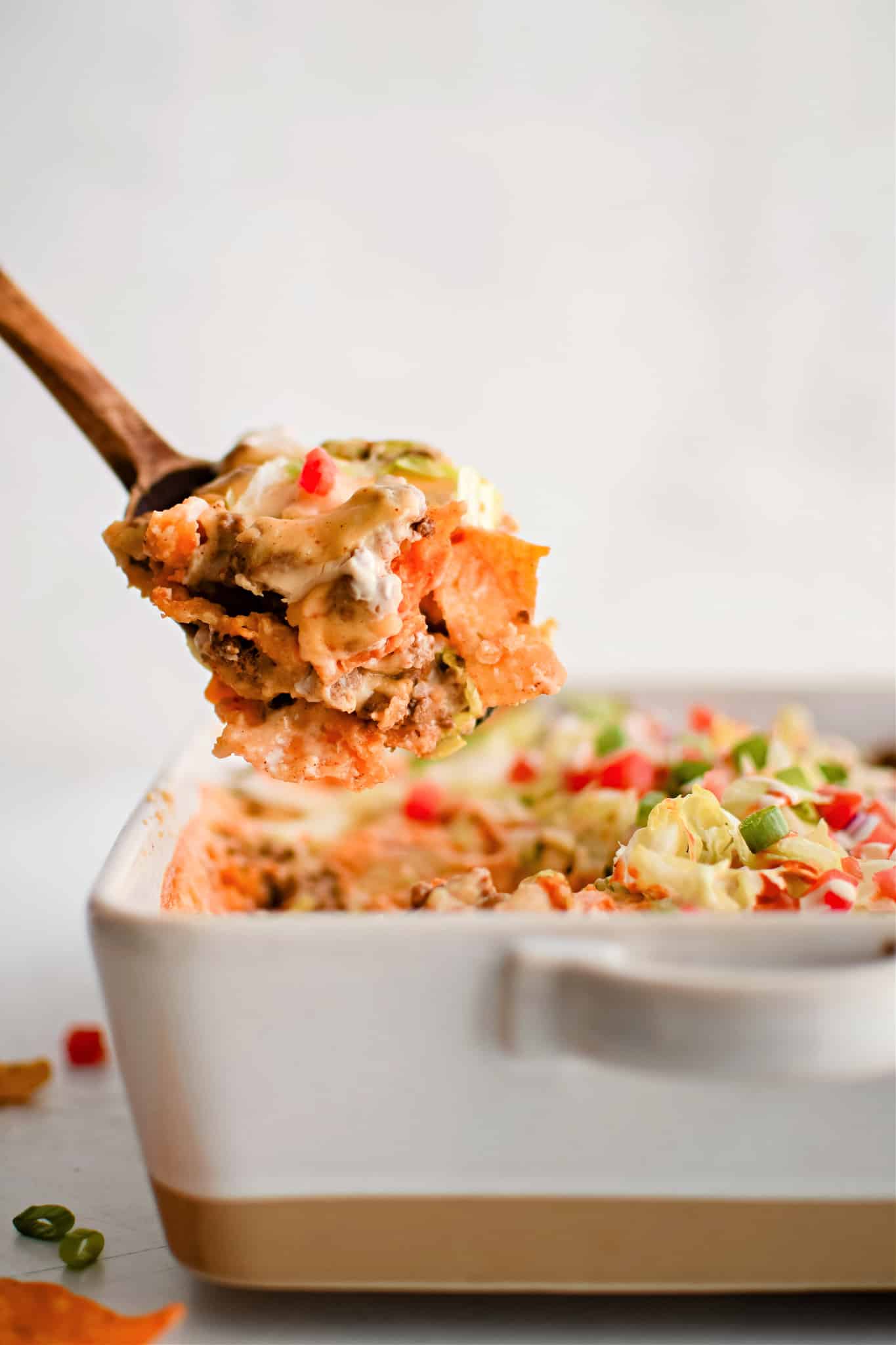 Wooden spatula raising a large portion of walking taco casserole from a white casserole pan filled with the remaining meat and cheese filled casserole.