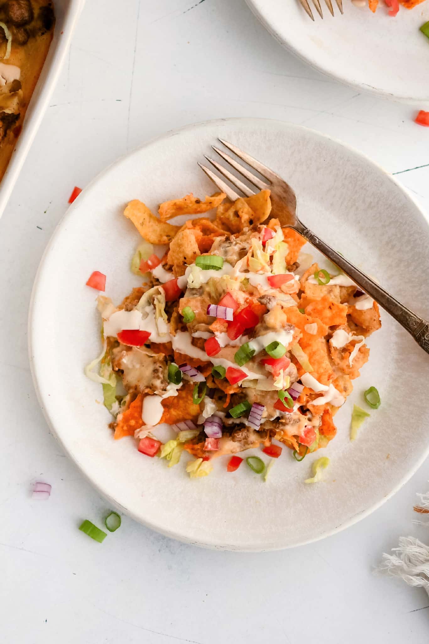 White dinner plate topped with a generous serving of walking taco casserole topped with shredded iceberg lettuce, chopped tomatoes, sour cream, and hot sauce.