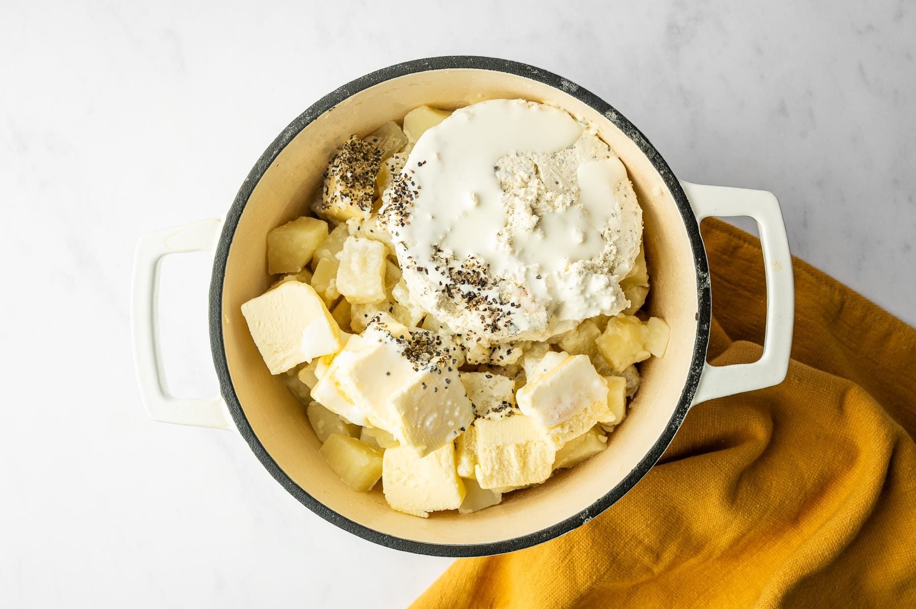 Cooked Russet potato chunks topped with pieces of salted butter, Boursin Garlic & Fine Herbs cheese, heavy cream, and cracked black pepper in a white enameled Dutch oven with a black rim, with a mustard-yellow towel underneath.