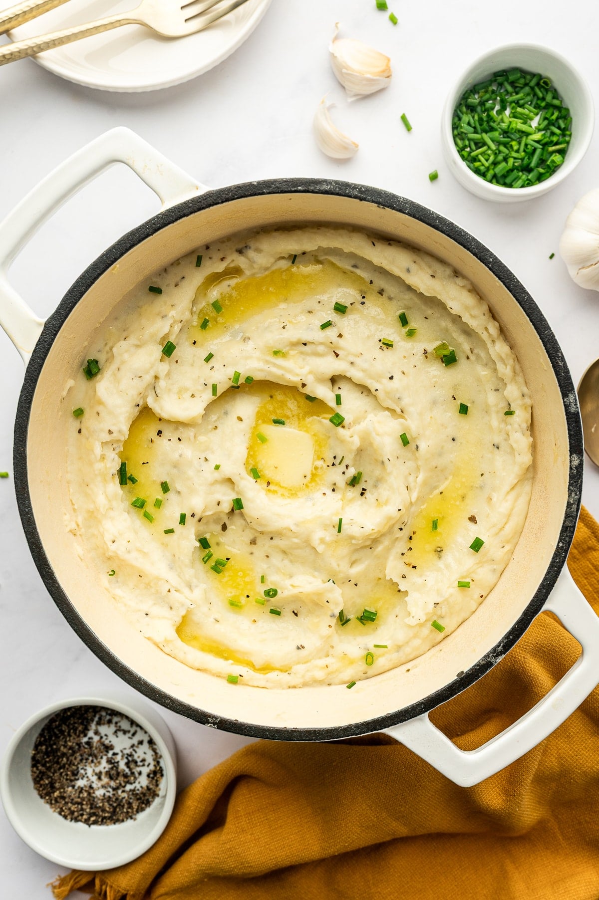 Creamy Boursin mashed potatoes swirled in a white enameled Dutch oven with a black rim and topped with melted butter, a pat of butter, chopped fresh chives, and black pepper, surrounded by garlic cloves, a small bowl of chives, a dish of pepper, and a mustard-yellow linen.