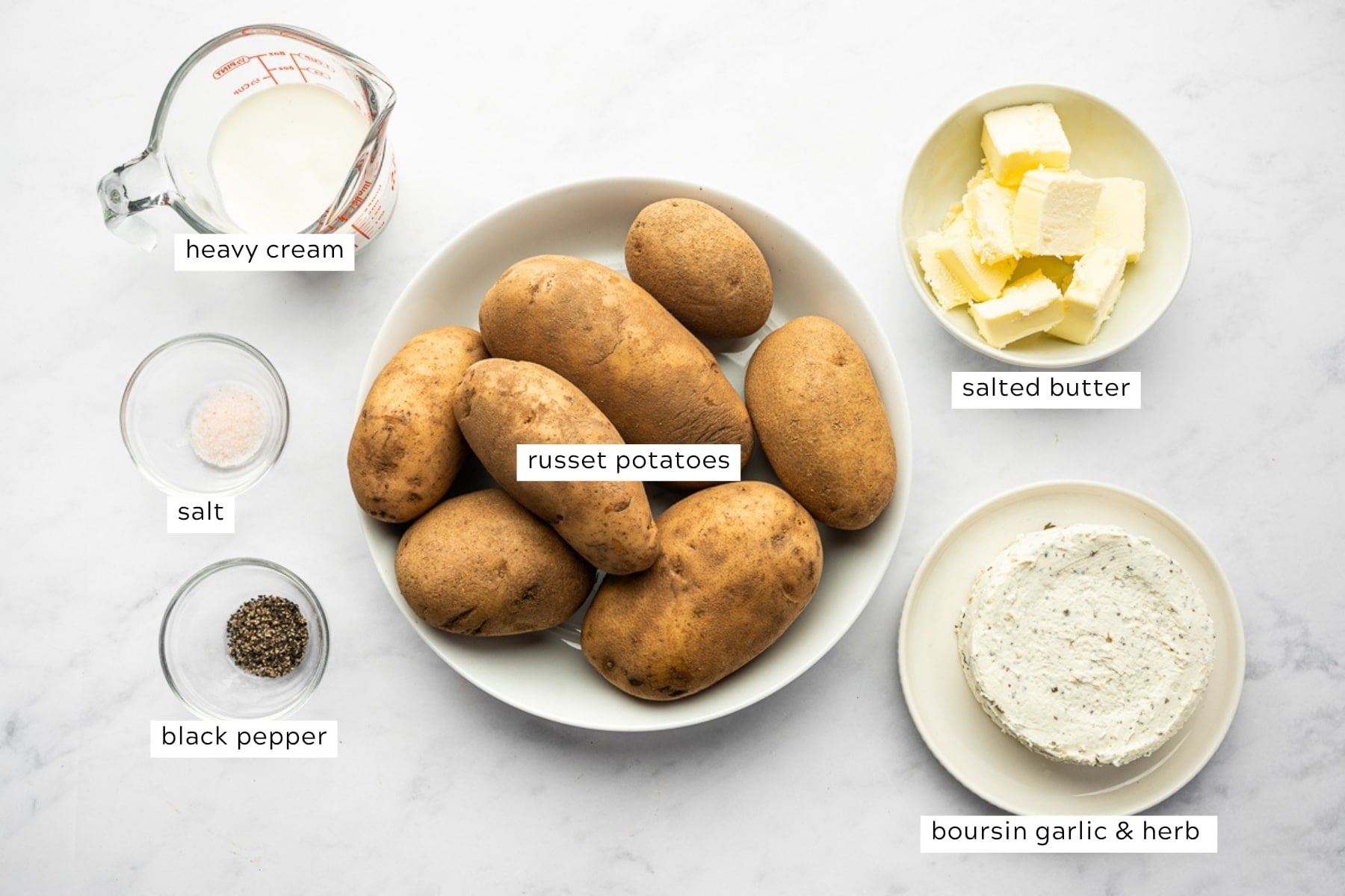 Overhead ingredient layout for Boursin mashed potatoes with whole Russet potatoes on a white plate, heavy cream in a glass measuring cup, cubed salted butter in a cream-colored bowl, Boursin Garlic & Fine Herbs cheese on a small plate, and separate bowls of salt and black pepper. Text overlays label each ingredient: "heavy cream," "russet potatoes," "salted butter," "boursin garlic & herb," "salt," and "black pepper."