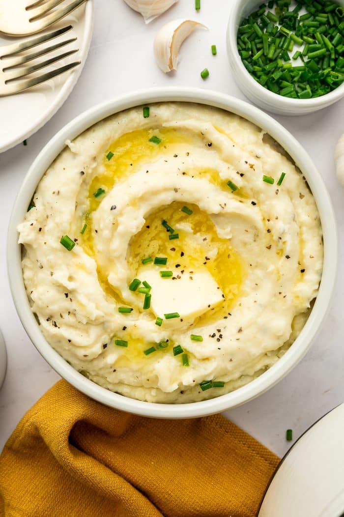 Creamy Boursin mashed potatoes in a round white serving bowl topped with a pat of butter, melted butter, chopped fresh chives, and black pepper, with a mustard-yellow linen, garlic cloves, gold forks, and a small white bowl of chopped chives surrounding the dish.