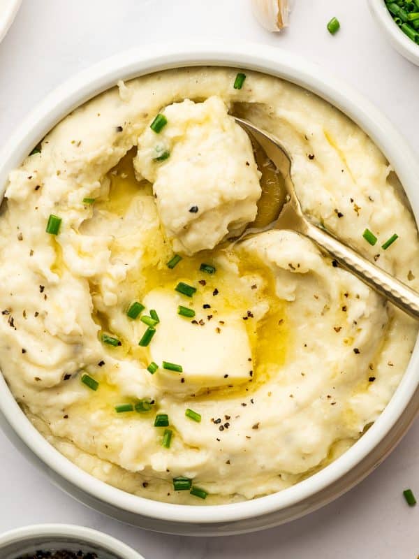 Close-up of creamy Boursin mashed potatoes in a white serving bowl with a gold spoon lifting a spoonful from the center, topped with melted butter, a pat of butter, chopped chives, and cracked black pepper.