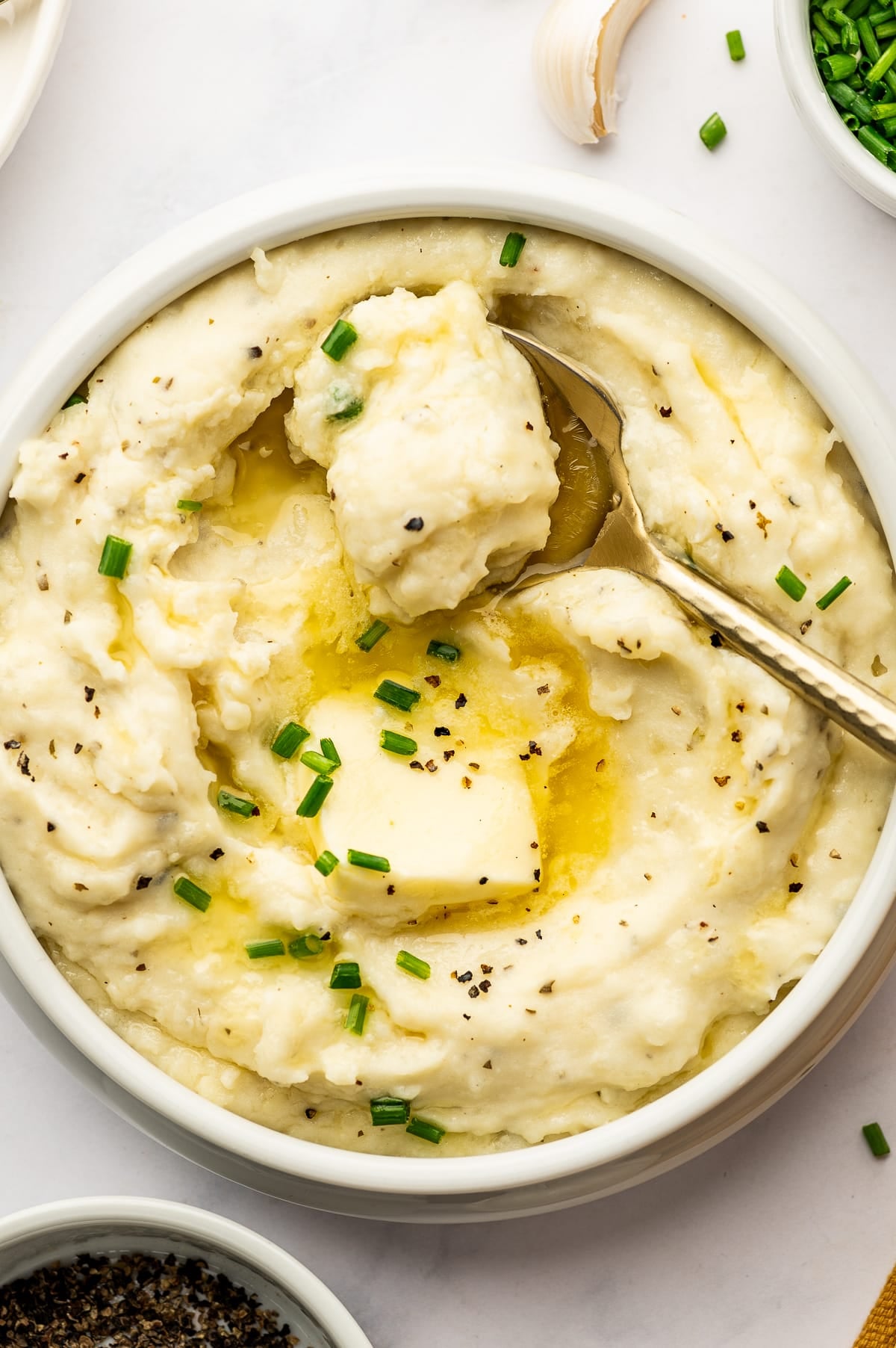 Close-up of creamy Boursin mashed potatoes in a white serving bowl with a gold spoon lifting a spoonful from the center, topped with melted butter, a pat of butter, chopped chives, and cracked black pepper.