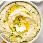Vertical Pinterest image showing creamy Boursin mashed potatoes in a white serving bowl topped with melted butter, a pat of butter, chopped chives, and black pepper. A large white text box across the top reads "Cheesy Boursin Mashed Potatoes," with "THEFORKEDSPOON.COM" printed along the bottom.