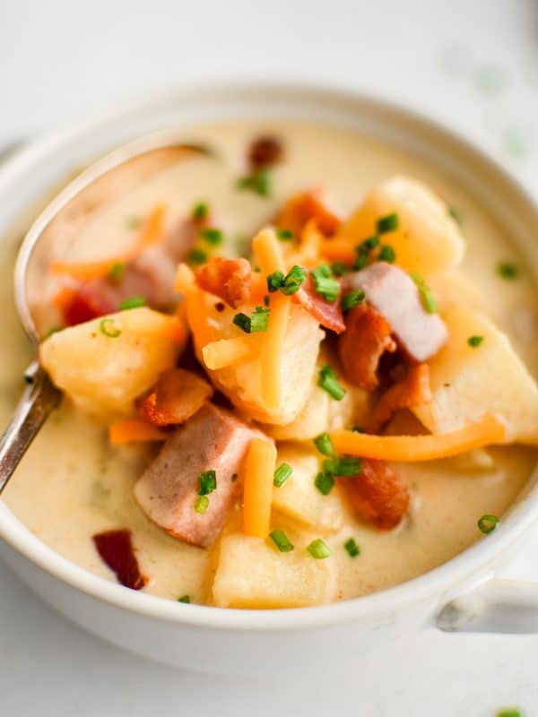 Close-up of a white handled bowl filled with creamy ham and potato soup, with a spoon resting inside. The surface shows large potato chunks, pieces of ham, shredded cheddar, crispy bacon bits, and chopped chives in a rich, pale creamy broth. No text overlay.