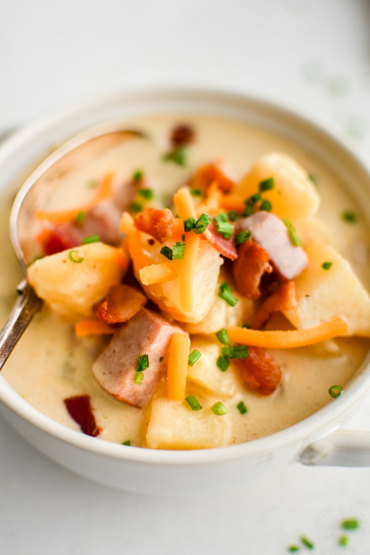 Close-up of a white handled bowl filled with creamy ham and potato soup, with a spoon resting inside. The surface shows large potato chunks, pieces of ham, shredded cheddar, crispy bacon bits, and chopped chives in a rich, pale creamy broth. No text overlay.