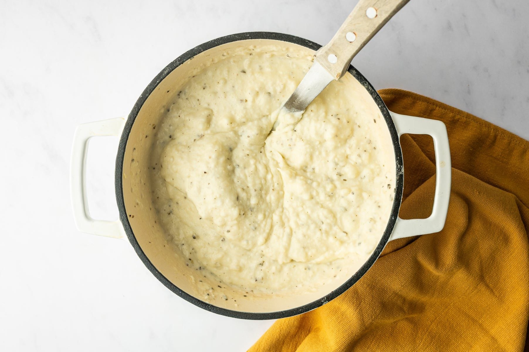 Creamy Boursin mashed potatoes with visible black pepper and herb flecks in a white enameled Dutch oven with a black rim, with a metal utensil resting in the potatoes and a mustard-yellow towel beneath the pot.