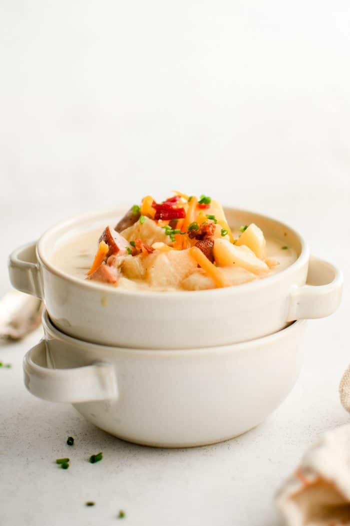 Two white handled soup bowls are stacked, with the top bowl filled with creamy ham and potato soup. The soup is garnished with shredded cheddar, chopped chives, and bacon bits, with chunky potatoes and ham visible near the top.