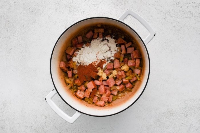 Overhead view of a white enamel Dutch oven on a light gray textured background with browned ham and softened vegetables in the bottom. A mound of white flour sits on the right side, paprika is sprinkled in the center, and minced garlic is added in small piles, showing the roux and seasoning step.
