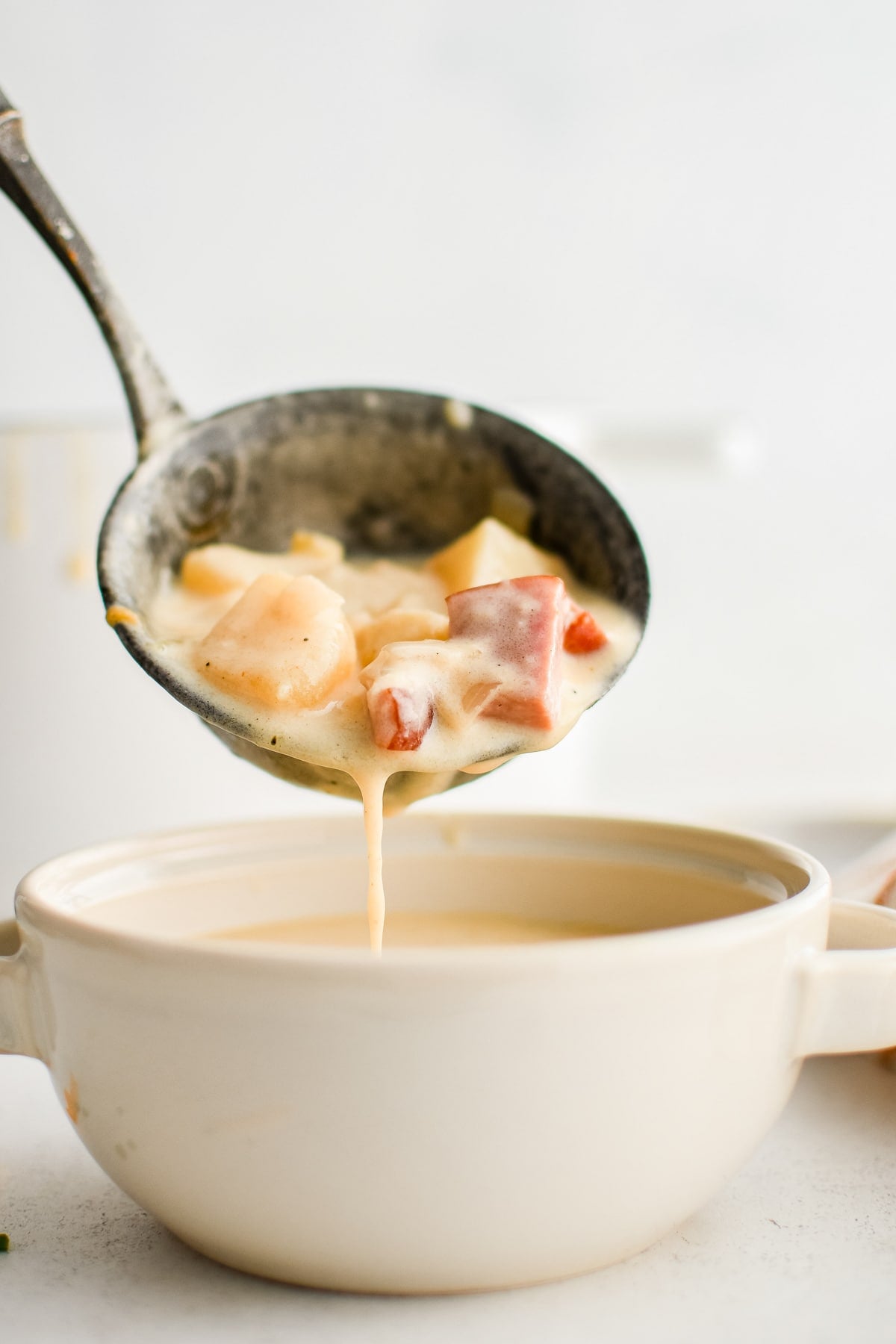 A metal ladle lifts a scoop of creamy ham and potato soup above a white handled soup bowl. Large potato chunks and pink ham pieces sit in the ladle, and the thick soup drips back toward the bowl against a bright, minimal background.