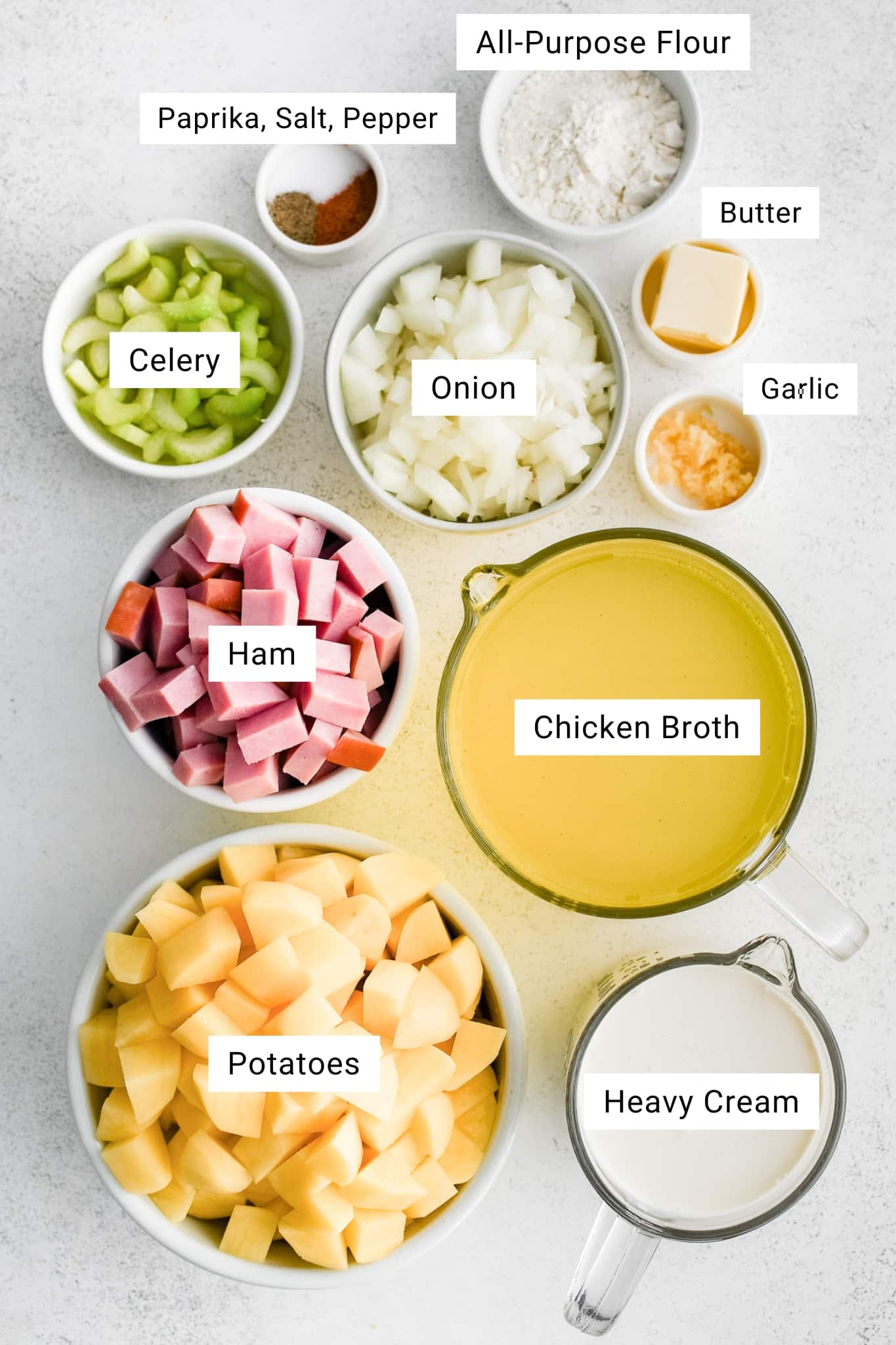 Overhead ingredients photo on a light gray stone surface with small bowls and measuring cups arranged neatly. White label text overlays identify each ingredient: “All-Purpose Flour,” “Paprika, Salt, Pepper,” “Butter,” “Garlic,” “Celery,” “Onion,” “Ham,” “Chicken Broth,” “Potatoes,” and “Heavy Cream.” No other text is present.