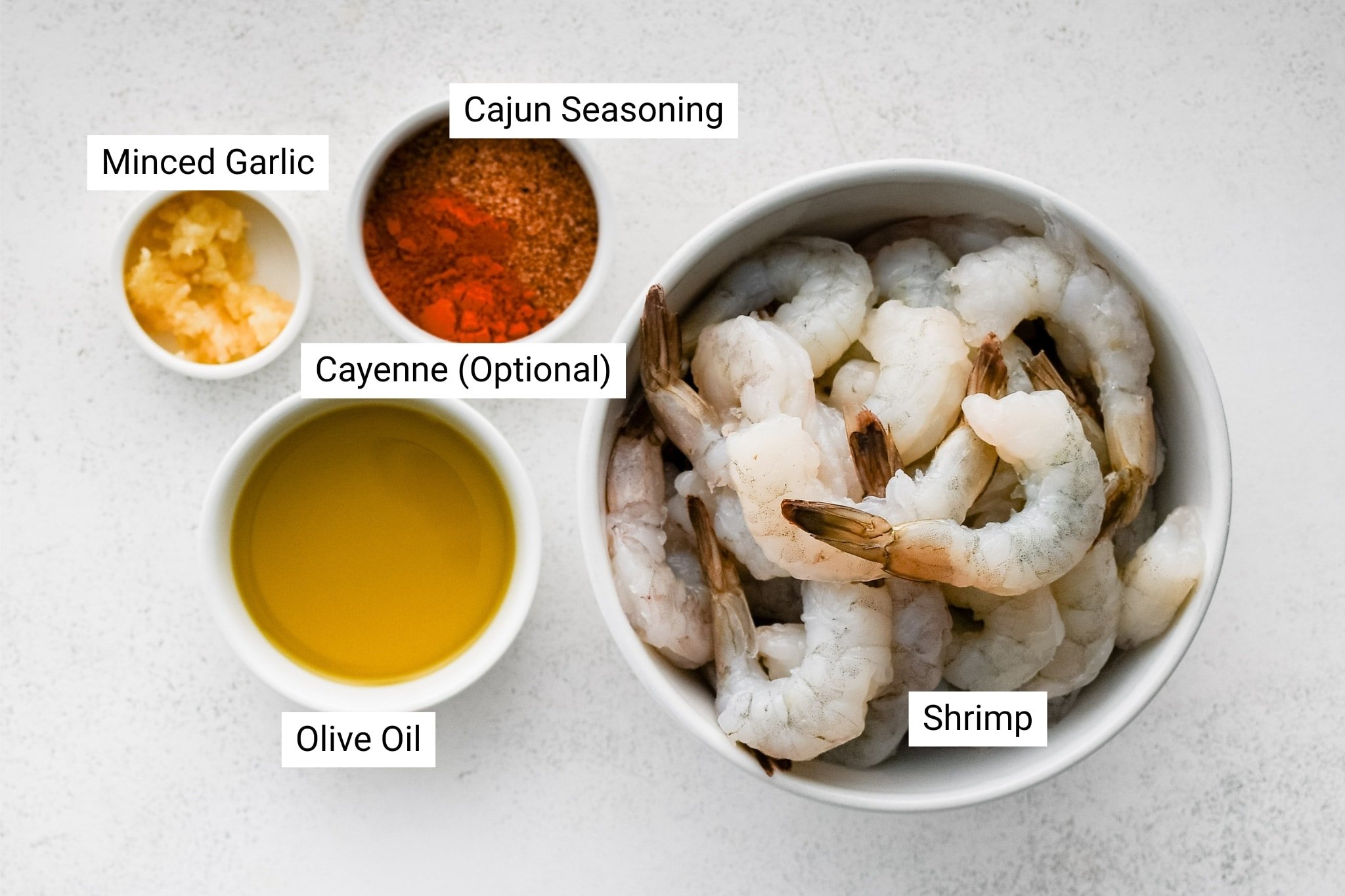 A large white bowl holds raw, peeled shrimp with tails on. Surrounding the shrimp are small bowls containing minced garlic, golden olive oil, a blend of Cajun seasoning, and a pinch of optional cayenne pepper for added heat. The ingredients are clearly labeled.