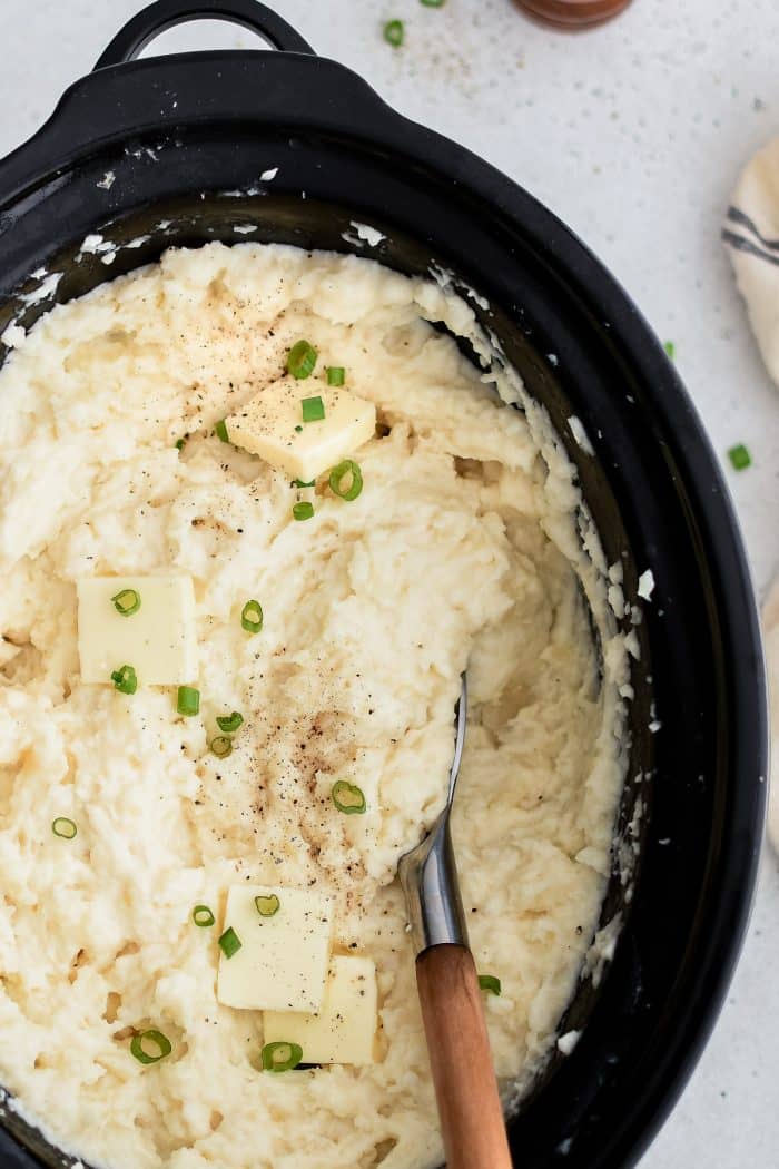 Large crockpot filled with buttery mashed potatoes garnished with green onions.