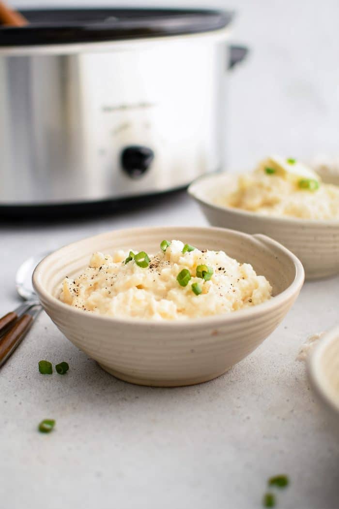 Bowl filled with creamy, buttery mashed potatoes garnished with green onions with a large crockpot in the back ground.