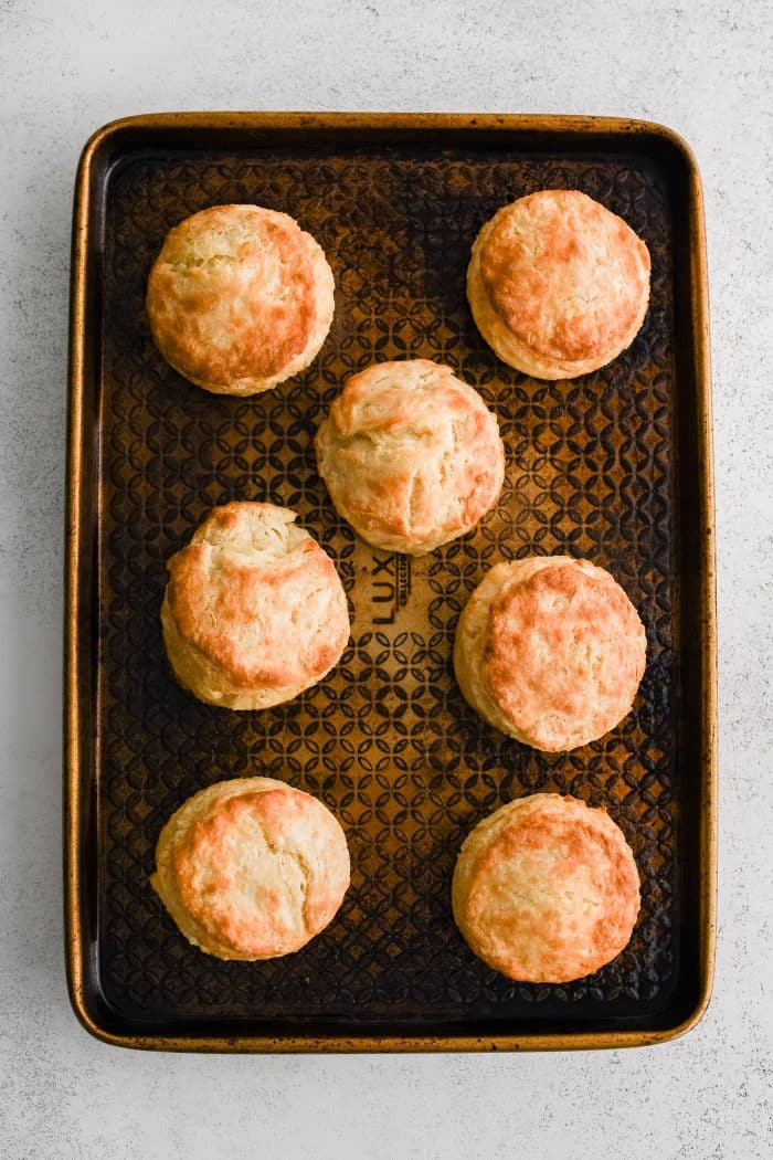 Seven perfectly golden, flakey, and buttery biscuits cooling on a large baking sheet.