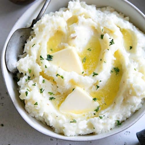 White bowl filled with creamy, rich, and fluffy mashed potatoes and topped with melted butter and minced parsley.