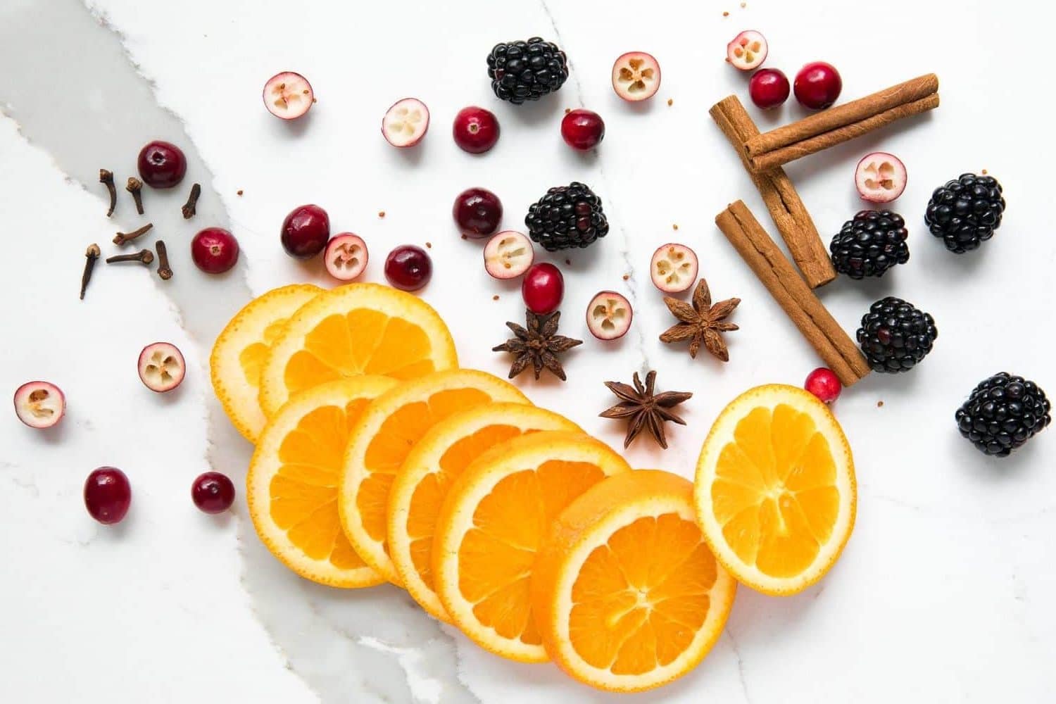 Overhead image of orange slices, whole cloves, cinnamon sticks, whole star anise, blackberries, and cranberries.